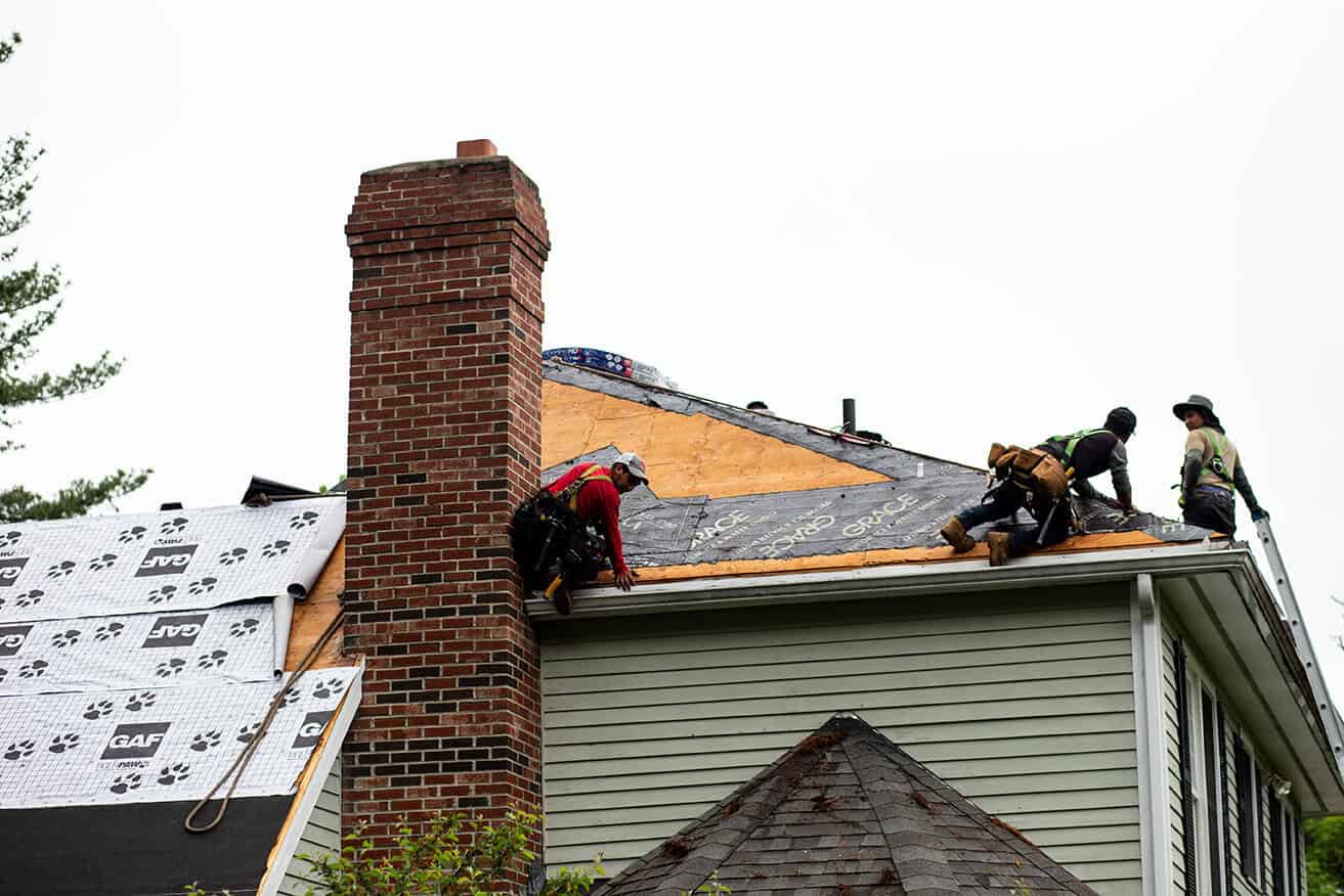 J. Carnes & Son Roofing crew in safety harnesses replacing a New Hampshire roof, with the old shingles stripped back to bare plywood decking and GAF underlayment and Grace ice and water shield being laid around the brick chimney.