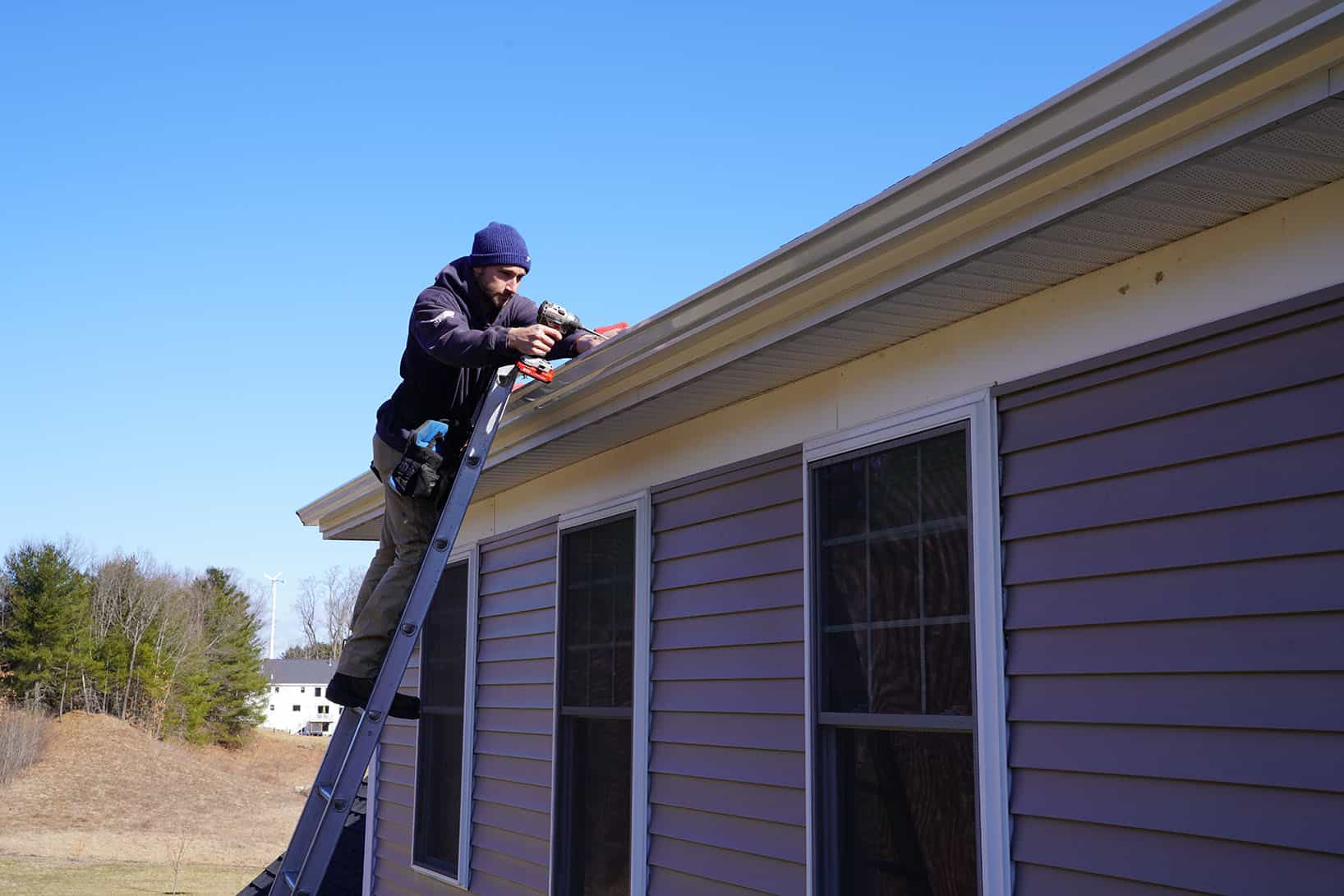 J. Carnes & Son Roofing crew member on a ladder fastening a white seamless gutter with a leaf guard to a gray clapboard New Hampshire home