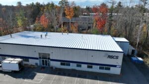 J. Carnes & Son Roofing crew finishing a white coated low-slope commercial roof on a single-story New Hampshire business, seen from above with fall foliage behind the building