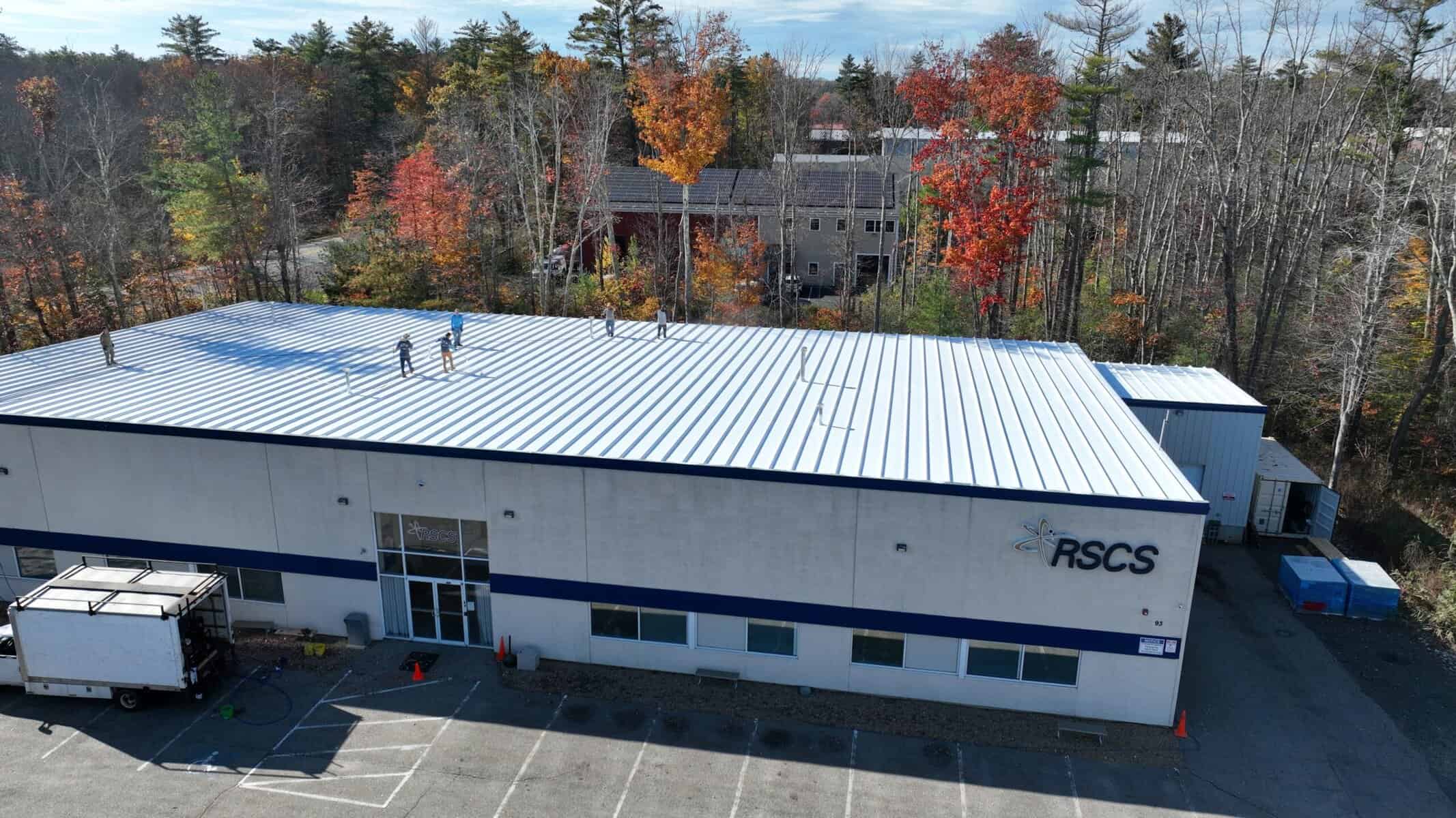 J. Carnes & Son Roofing crew finishing a white coated low-slope commercial roof on a single-story New Hampshire business, seen from above with fall foliage behind the building
