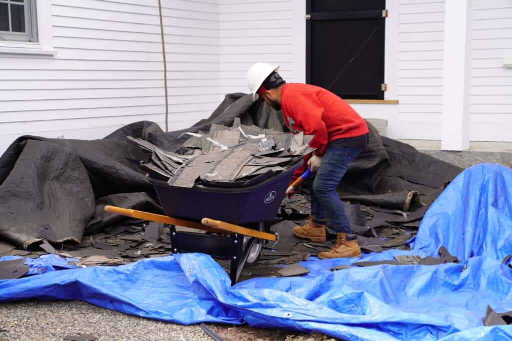 J. Carnes & Son Roofing crew member in a hard hat clearing torn-off shingle debris into a wheelbarrow over protective tarps during a roof repair on a New Hampshire Seacoast home