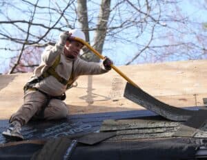 J. Carnes & Son Roofing crew member in a hard hat and safety harness tearing off worn asphalt shingles during a roof replacement on the New Hampshire Seacoast